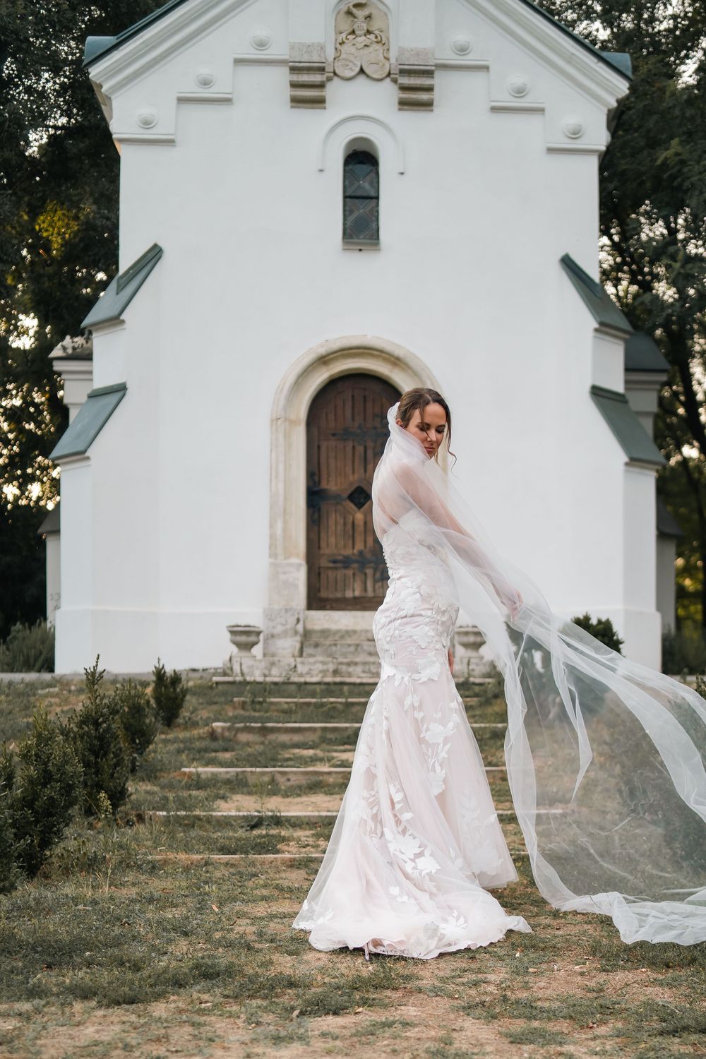 A bride in a white lace dress poses with flowing veil outside a charming white chapel during a rustic countryside wedding.