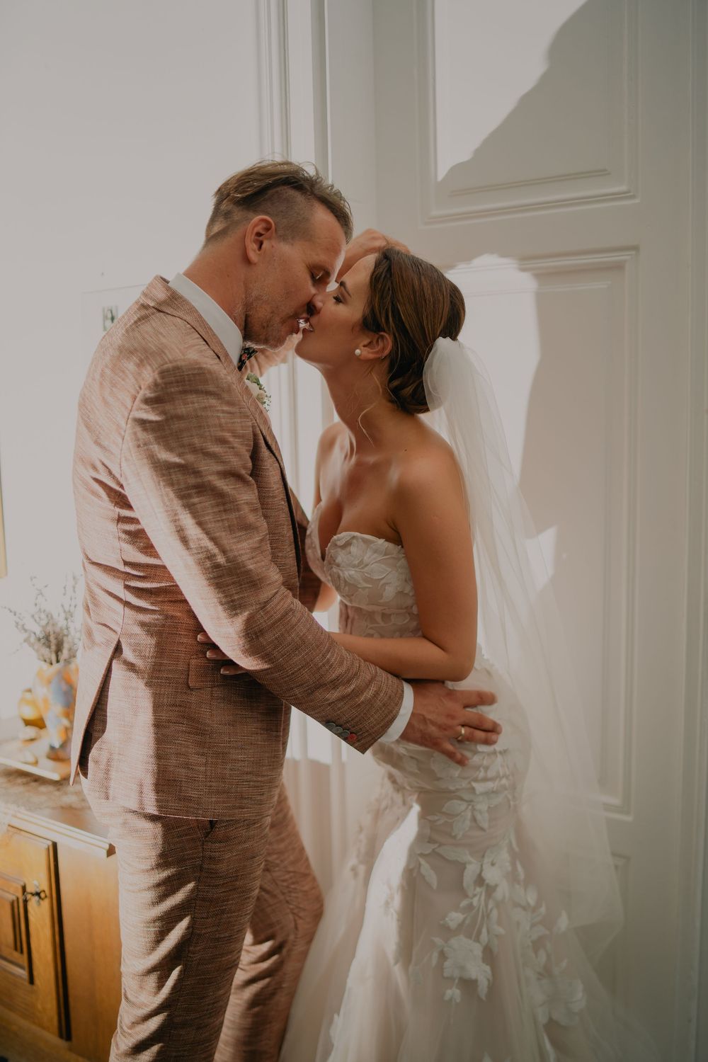 Bride in white dress and groom in tan suit share romantic kisses in a softly lit room during intimate wedding moments.