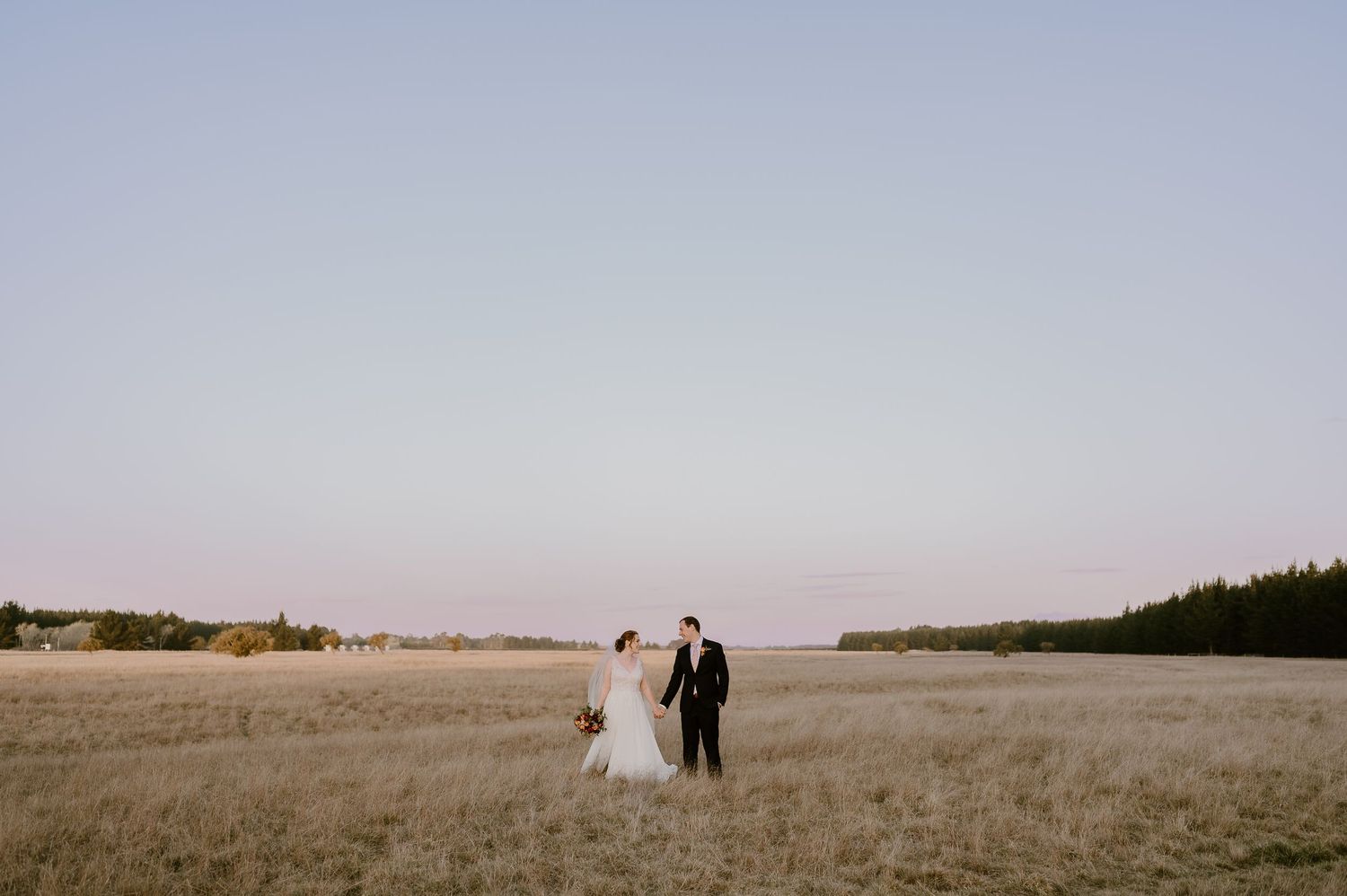 A couple walks away from the camera through a golden field at sunset with a distant treeline on the horizon.