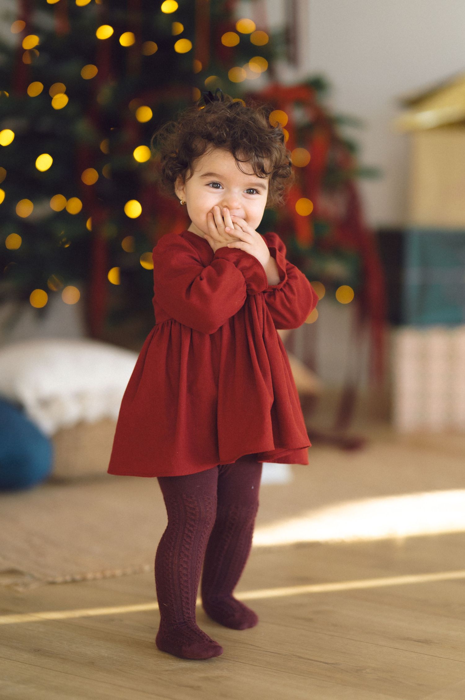 Small figure in red dress and burgundy tights sits on wood floor with festive Christmas lights twinkling in background.