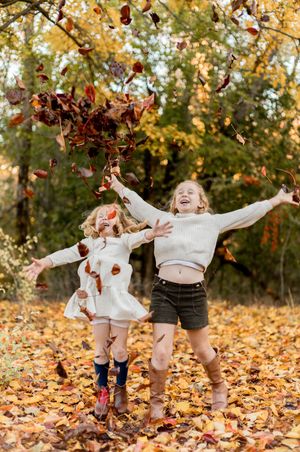 Two children playing in a pile of colorful autumn leaves beneath trees on a fall day.
