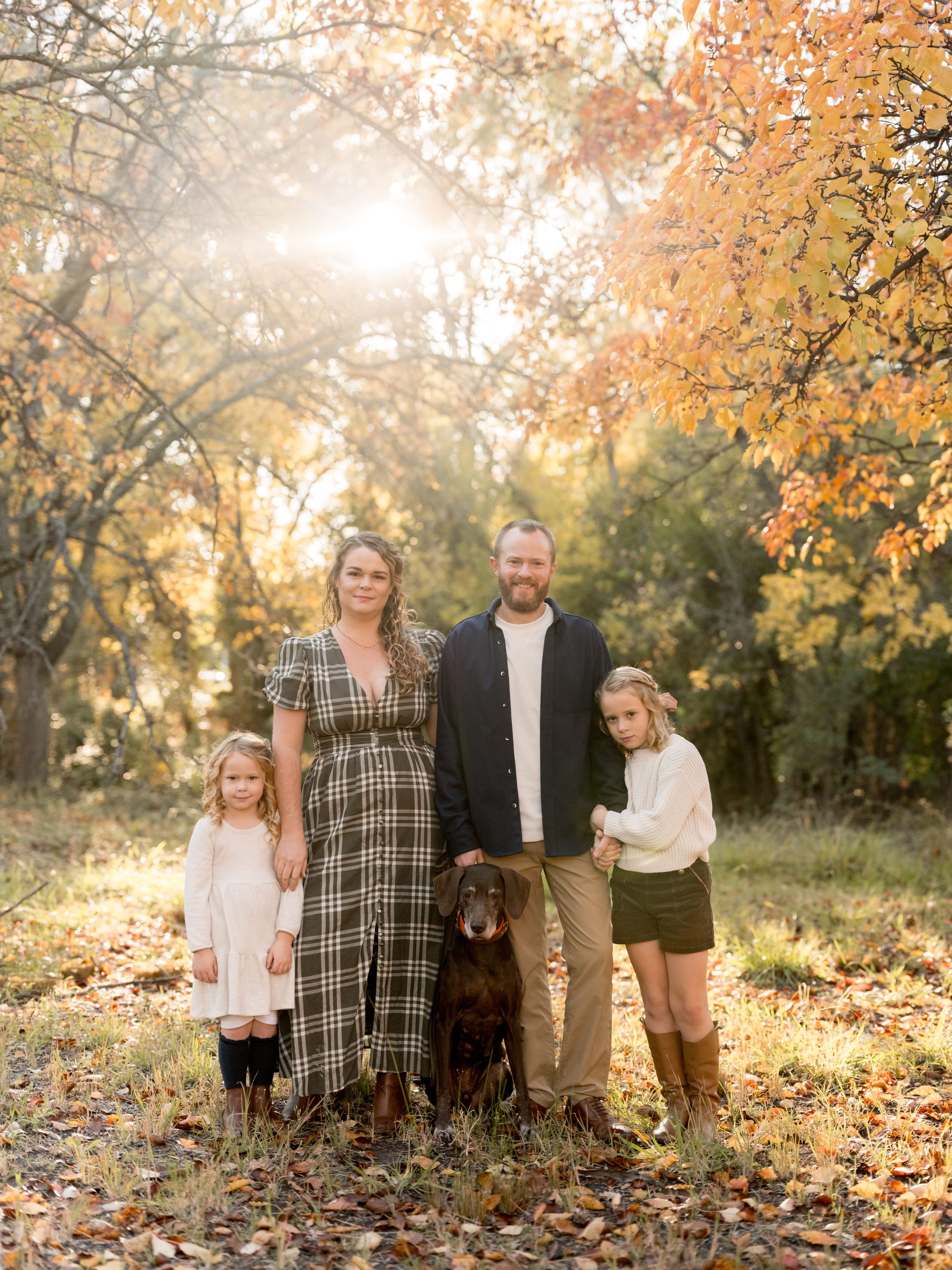A family gathers for outdoor autumn photos among fallen leaves and golden sunlight streaming through trees.
