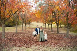 A couple stands between autumn trees with fallen red leaves and luggage beside them on a foggy fall morning.