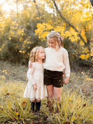 Two young children stand together outdoors on an autumn day with golden foliage in the background.