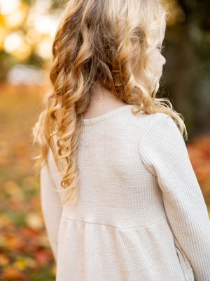 A person with wavy blonde hair wearing a cream colored knit sweater against a blurred autumn background.
