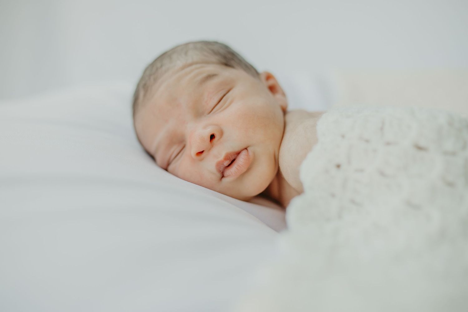 A peaceful newborn baby sleeping soundly on white bedding in soft natural light.