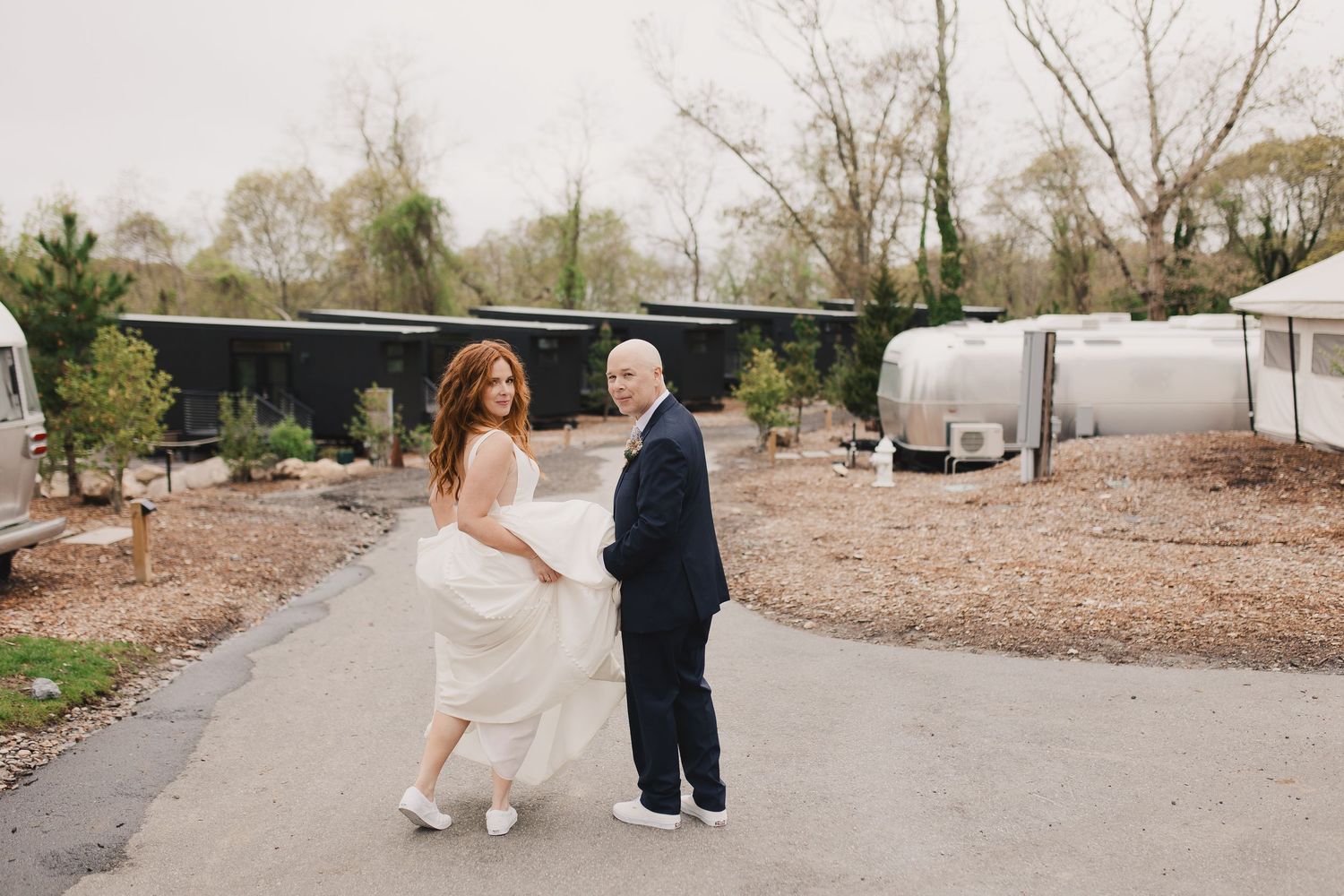 Bride in white gown and groom in navy suit walk together on a path surrounded by Airstream trailers outdoors.