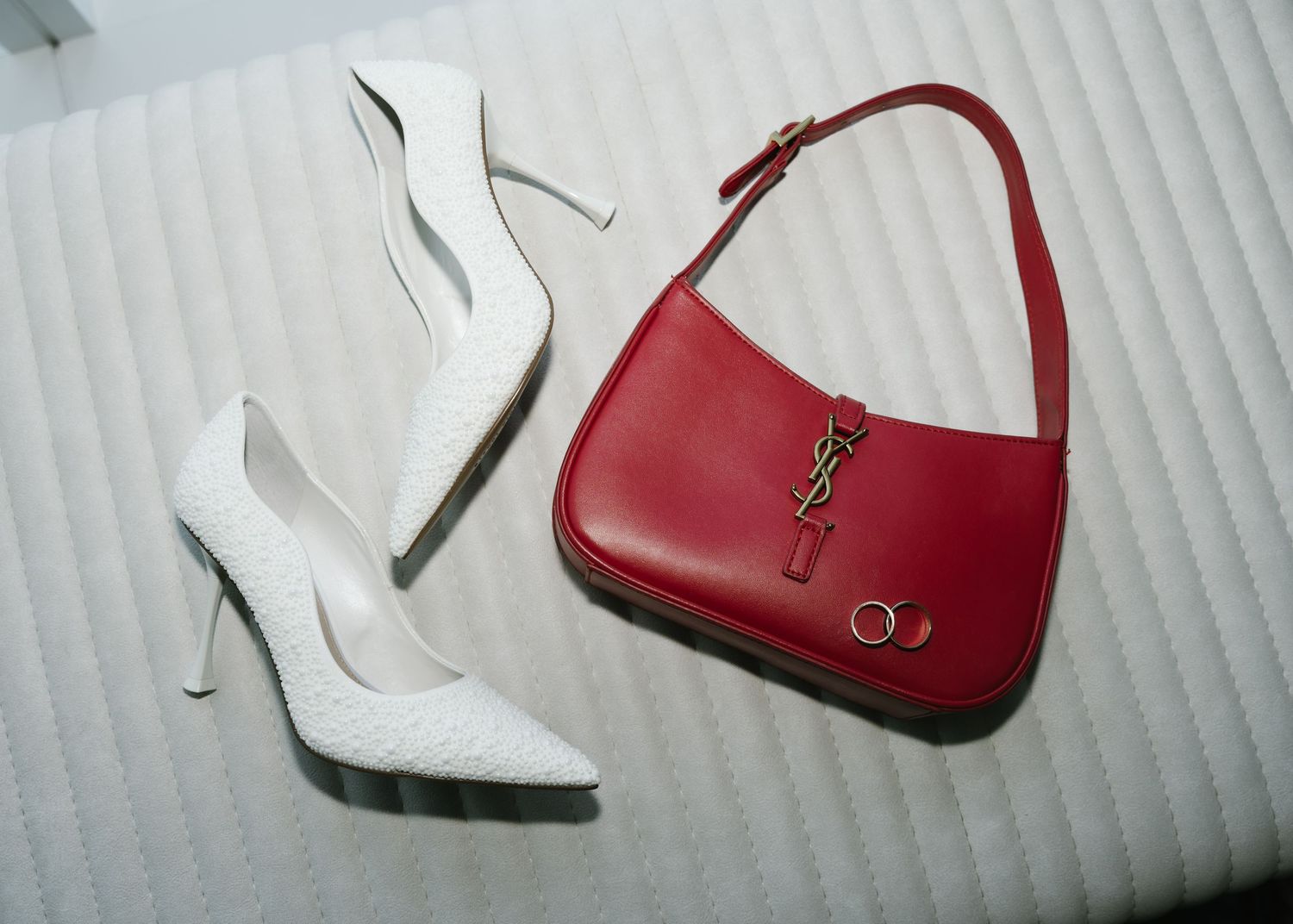 A pair of white stiletto heels next to a red YSL leather handbag on a textured white surface.
