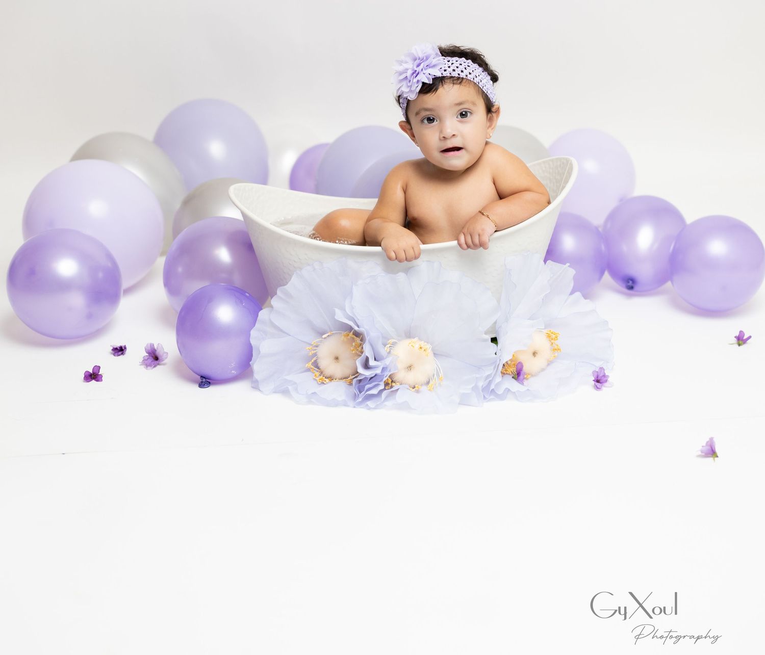A baby sits in a bubble bath surrounded by purple balloons for a milestone photography session.