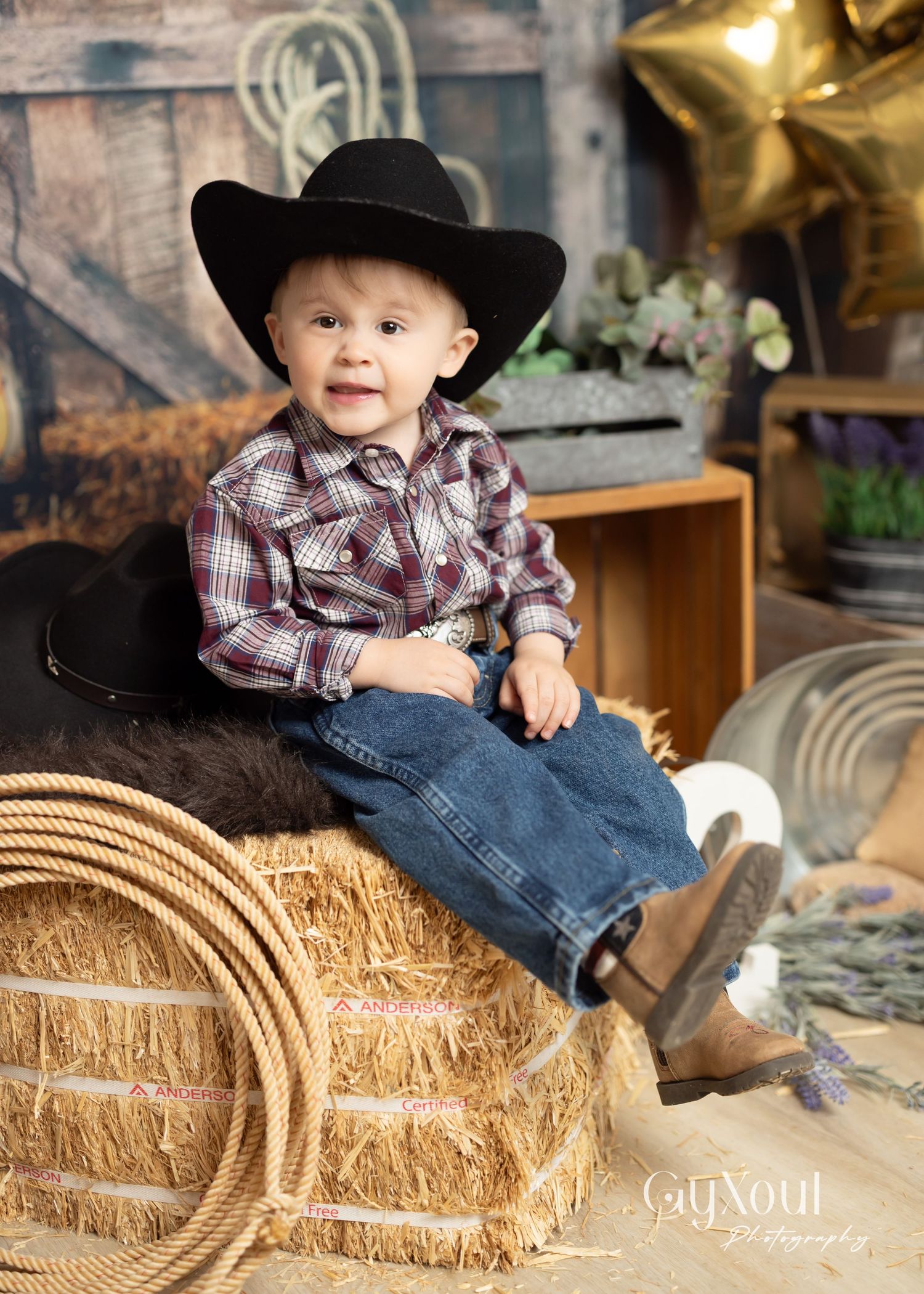 Little one in cowboy attire sits on hay bale with rope and rustic barn backdrop for western themed photoshoot.