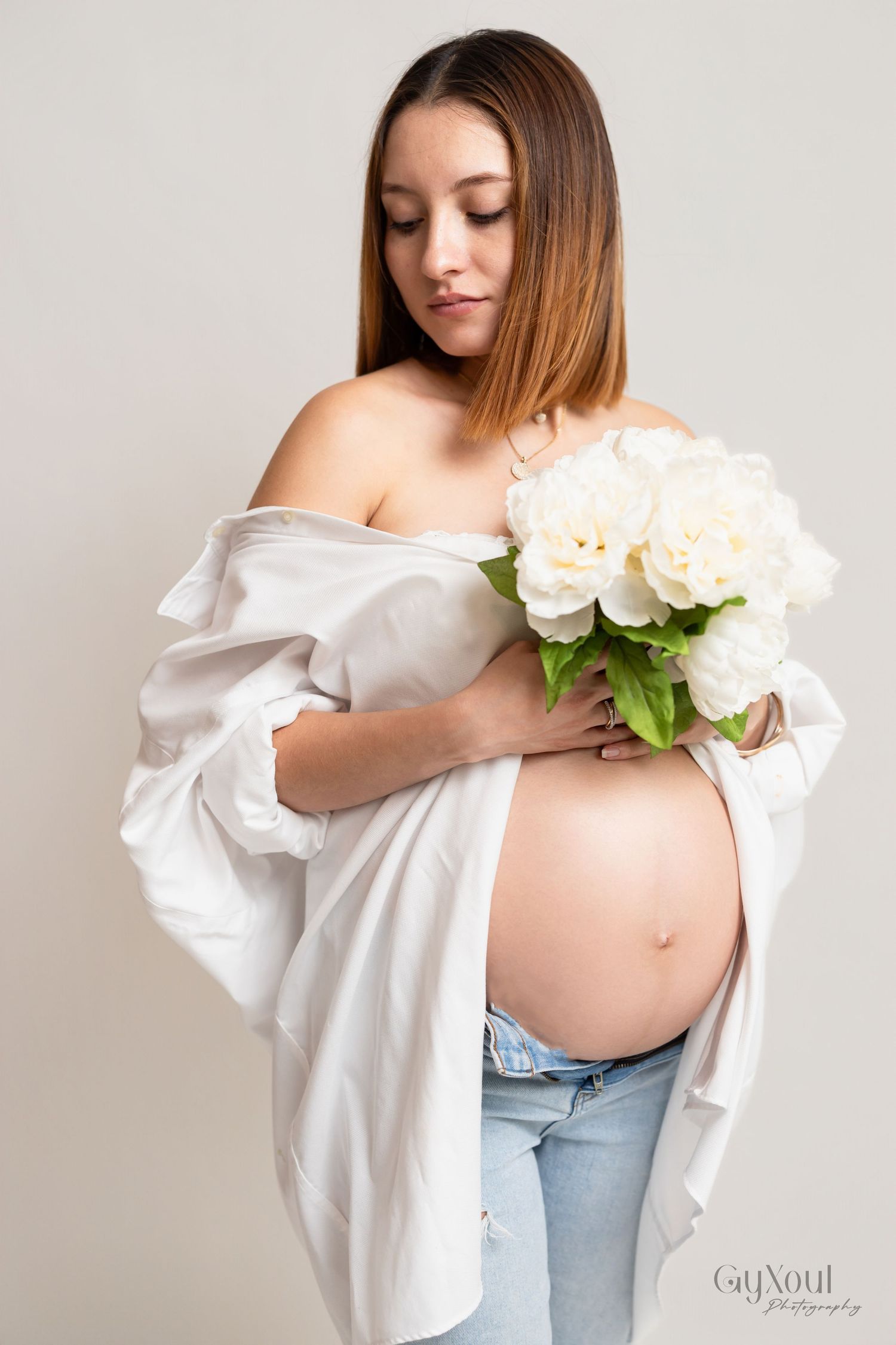 A pregnant woman in jeans and white shirt holds soft peach roses against her baby bump in an elegant maternity photo.