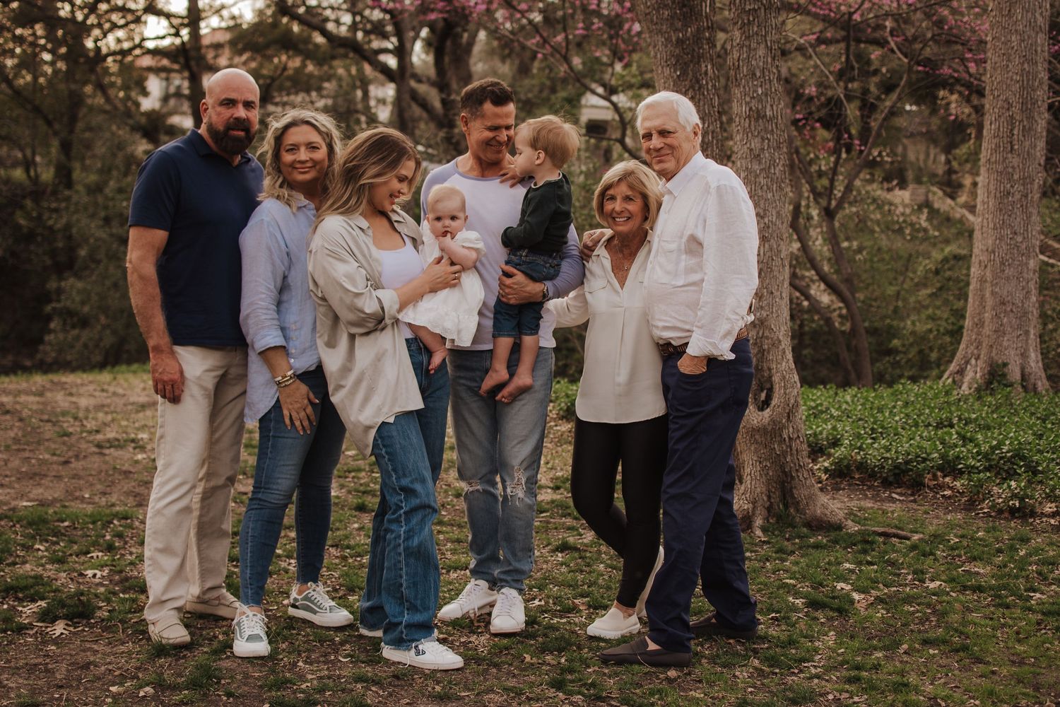 A multi-generational family gathers outdoors for a casual portrait in a wooded setting during autumn.