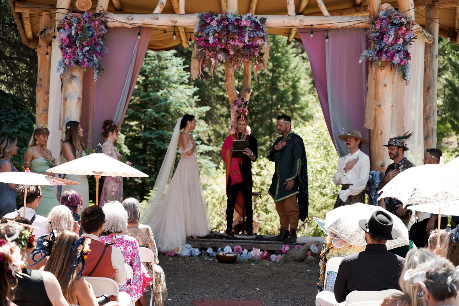 Wedding ceremony takes place under a wooden gazebo decorated with purple flowers and draped fabric.