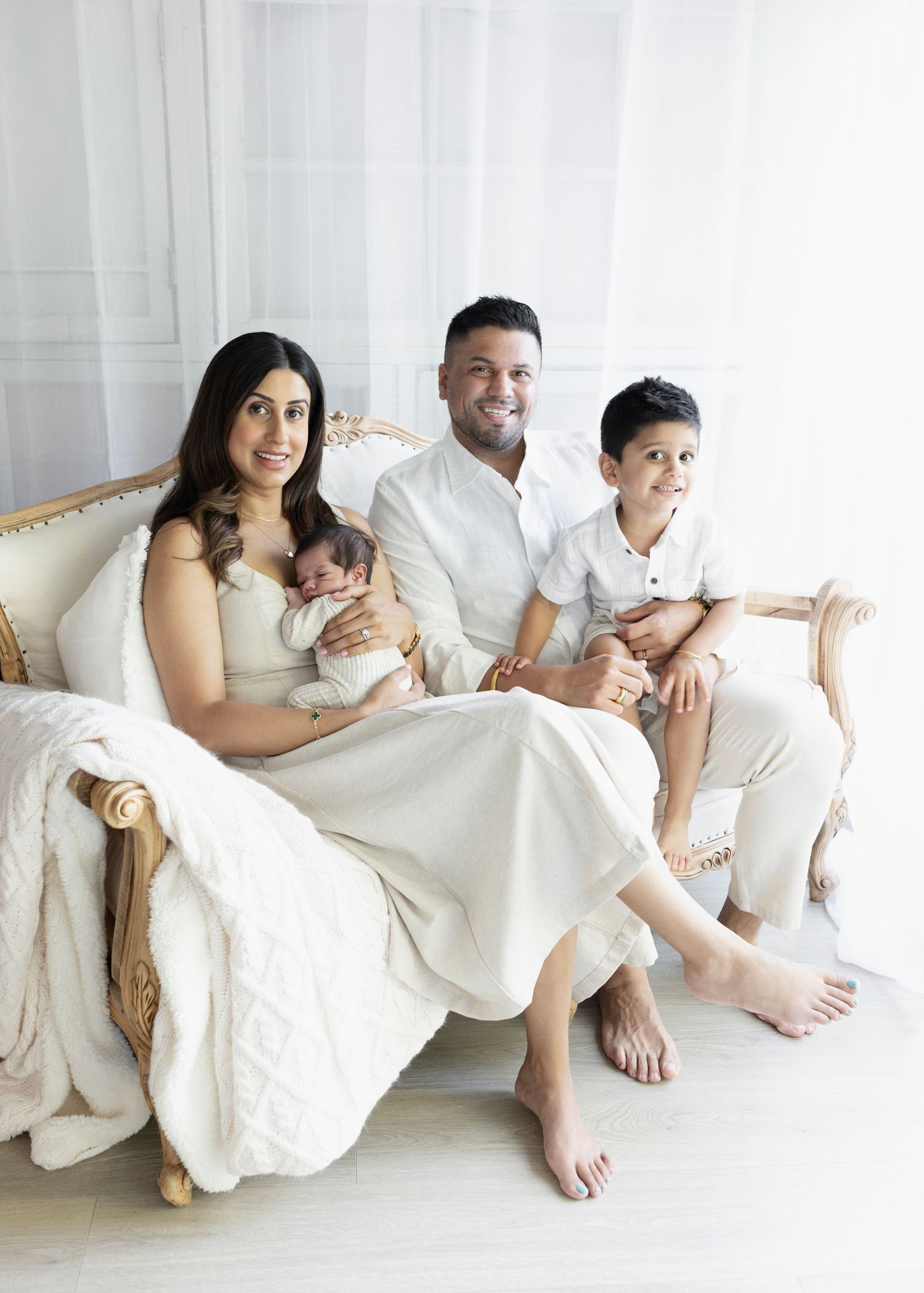 mom and dad sit on a white sofa in an all white setup with their newborn son and older son. Elizabeth Lang Photography