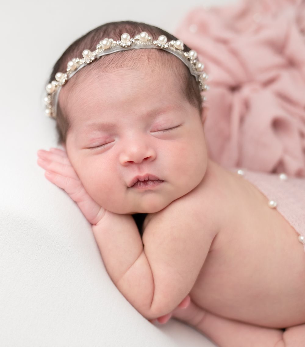 A sleeping newborn baby wearing a delicate lace headband rests on pink fabric in a professional portrait setting.