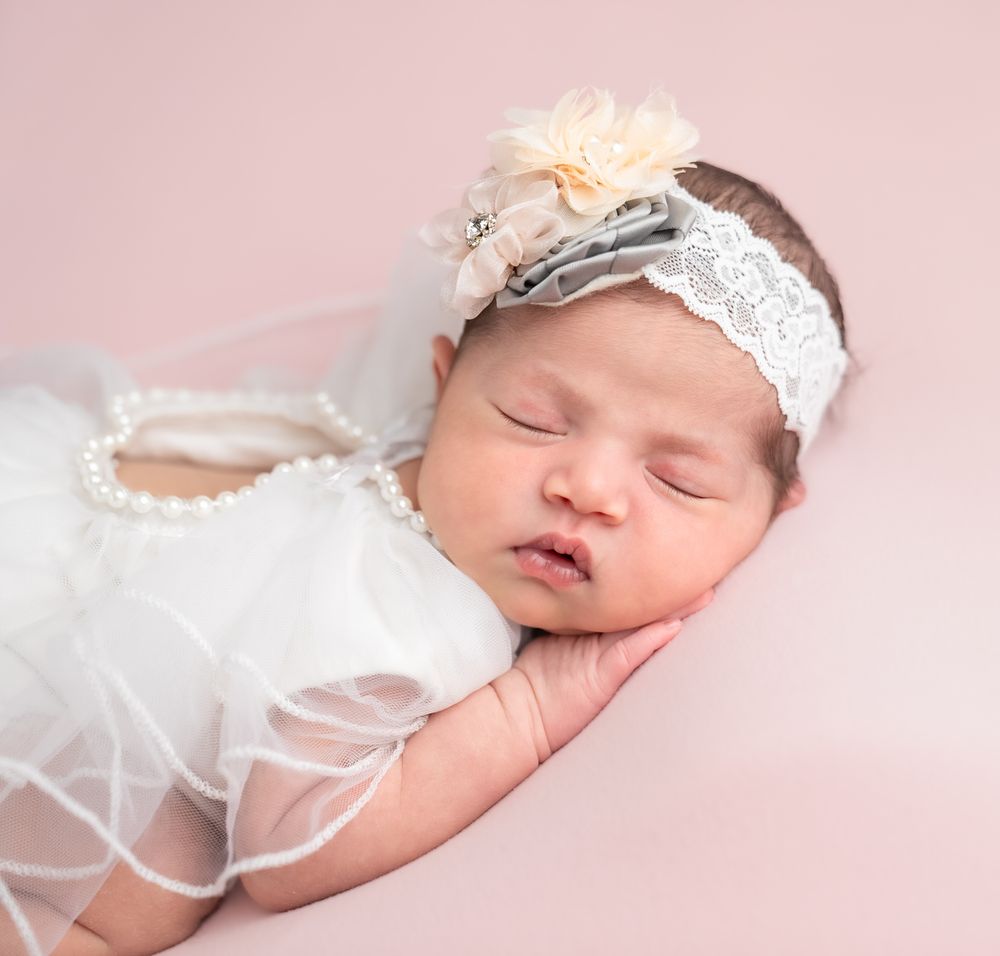 A sleeping newborn wearing a white lace dress and delicate floral headband lying on pink fabric.
