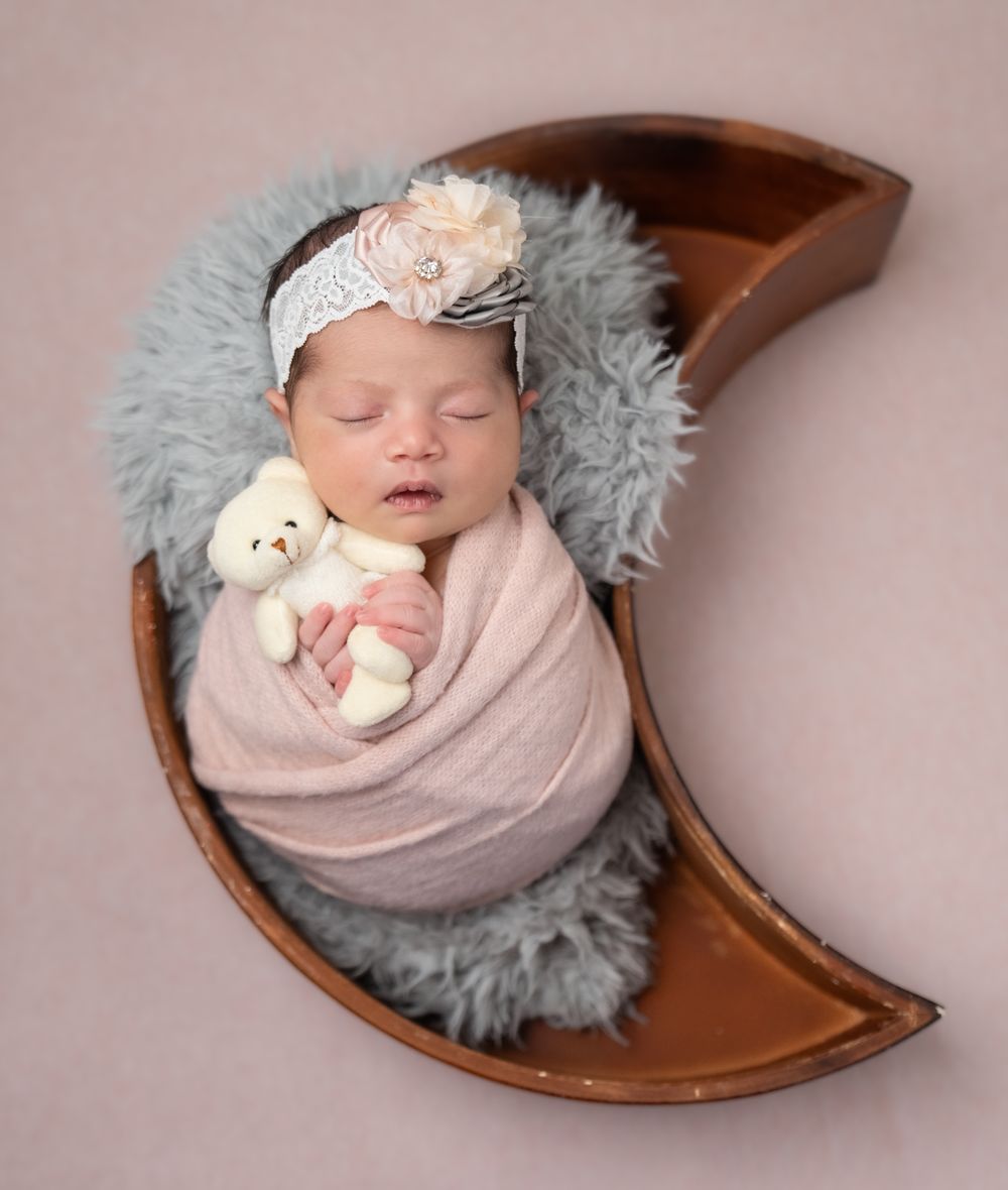 Sleeping newborn swaddled in pink wrap with lace headband rests in crescent moon prop on gray fur.