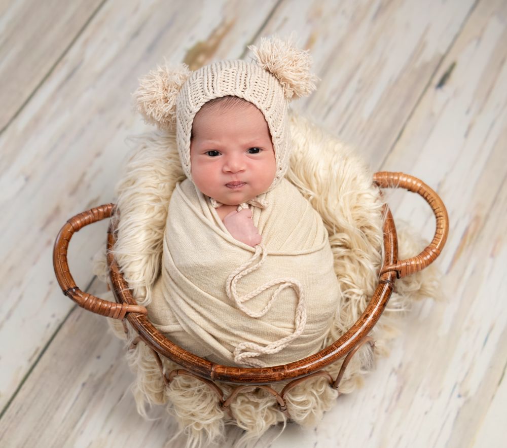 A baby in a cream wrap lies in a woven basket with fur lining while wearing a knit bear ear bonnet.