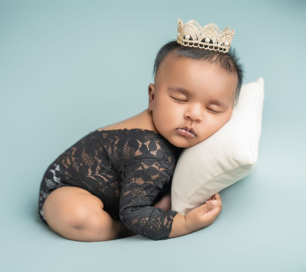 A sleeping infant wearing a crown and lace outfit rests peacefully against a white pillow on a blue background.
