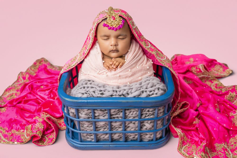 Newborn wrapped in pink and gold sari fabric rests in blue plastic basket during cultural portrait session.