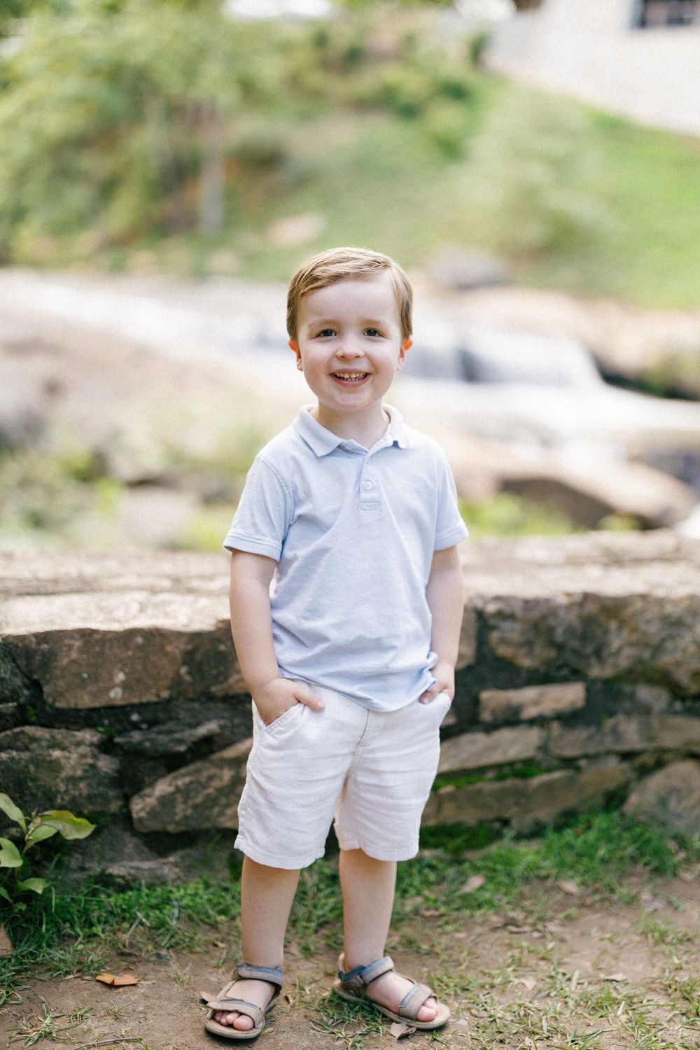A young child stands smiling outdoors wearing a light blue polo shirt, white shorts and sandals near a stone wall.