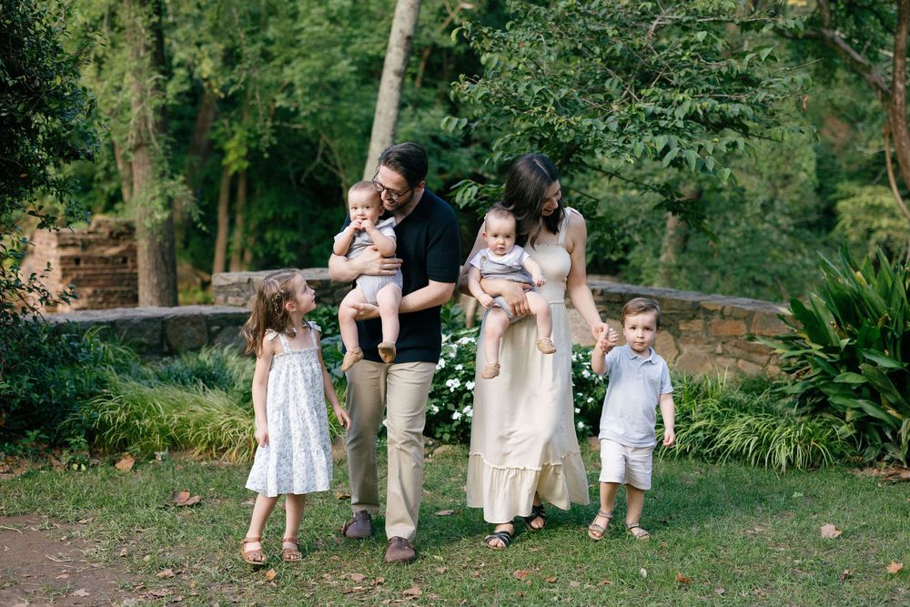 Extended family sits together outdoors on stone wall during summer photo session in lush garden setting.
