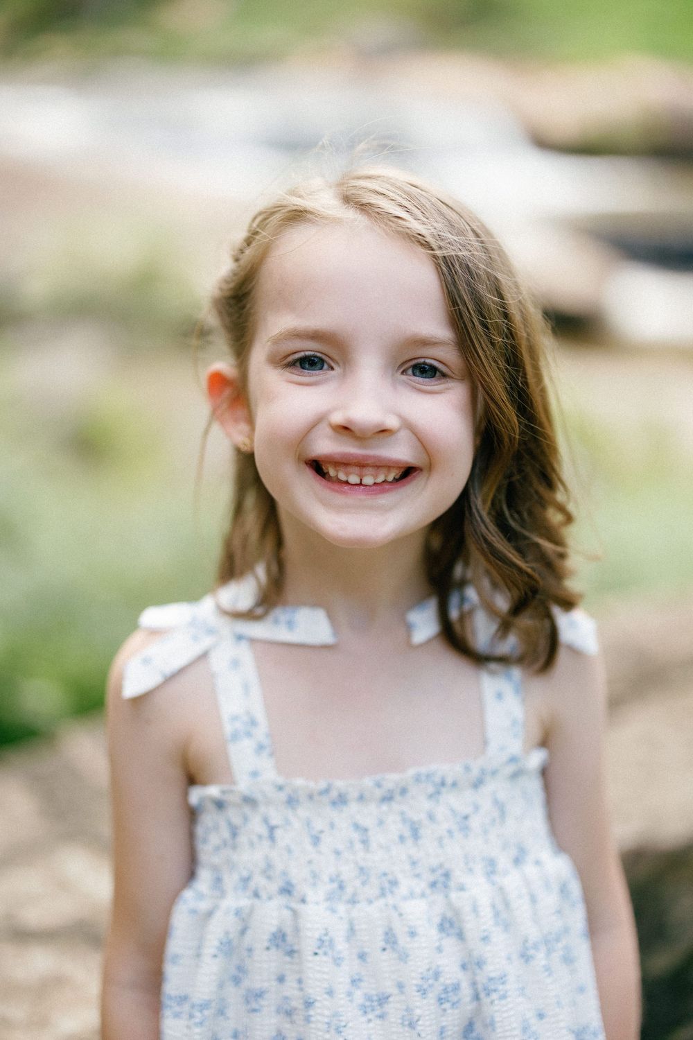 A young child in a floral summer dress smiles brightly at the camera in an outdoor setting.