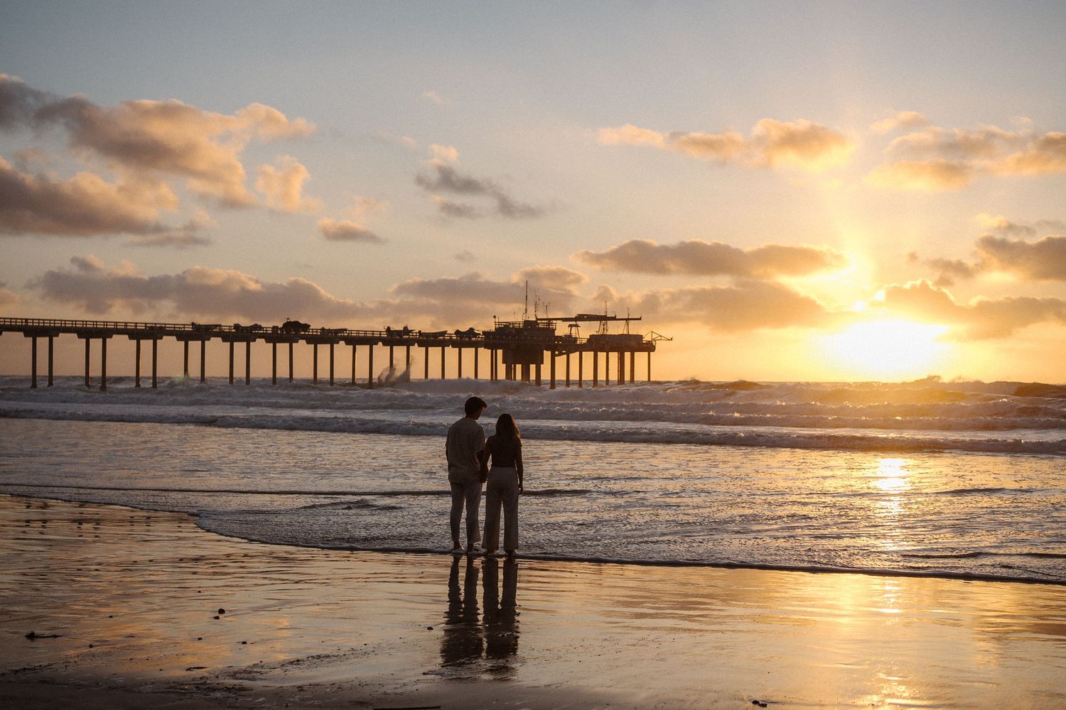 A silhouetted couple stands on a beach at sunset with a wooden pier and vibrant orange sky in La Jolla, California.
