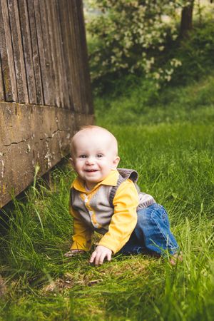 Baby in yellow sweater crawling on green grass near wooden fence post.