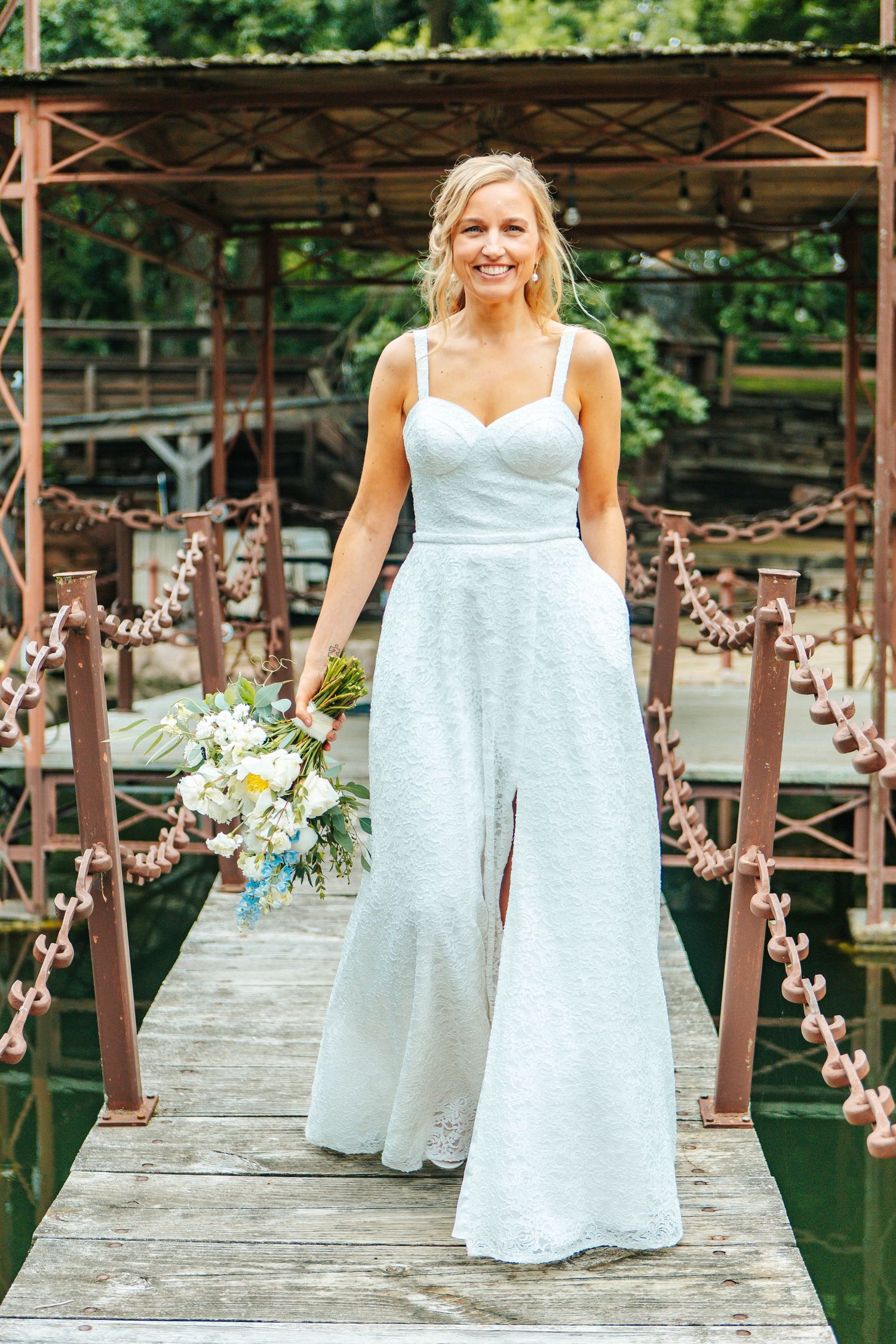 A bride in a white lace jumpsuit stands on a wooden bridge decorated with floral garlands and rustic railings.