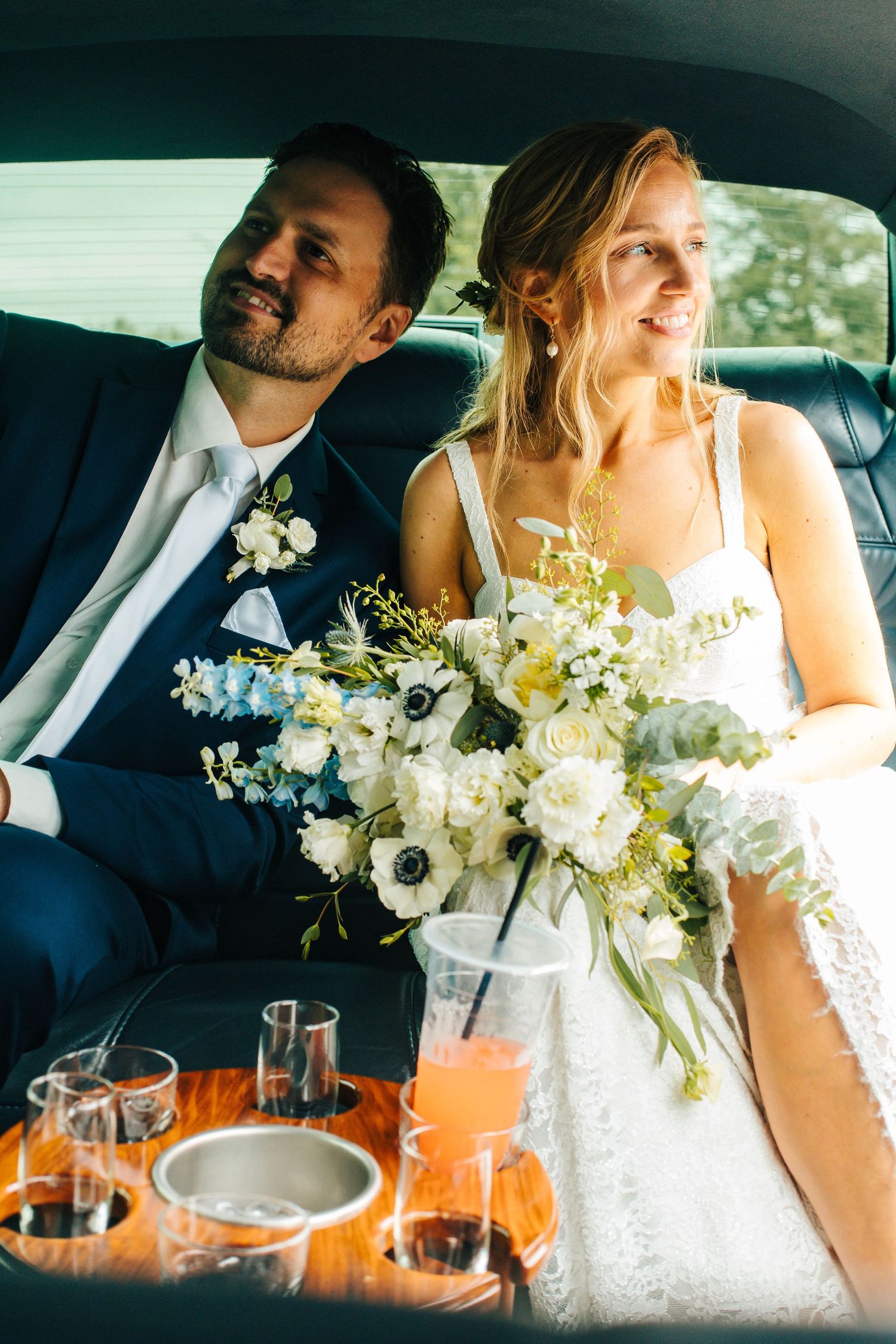 Newlyweds sit in back of car with champagne and beautiful white floral bouquet on wedding day.