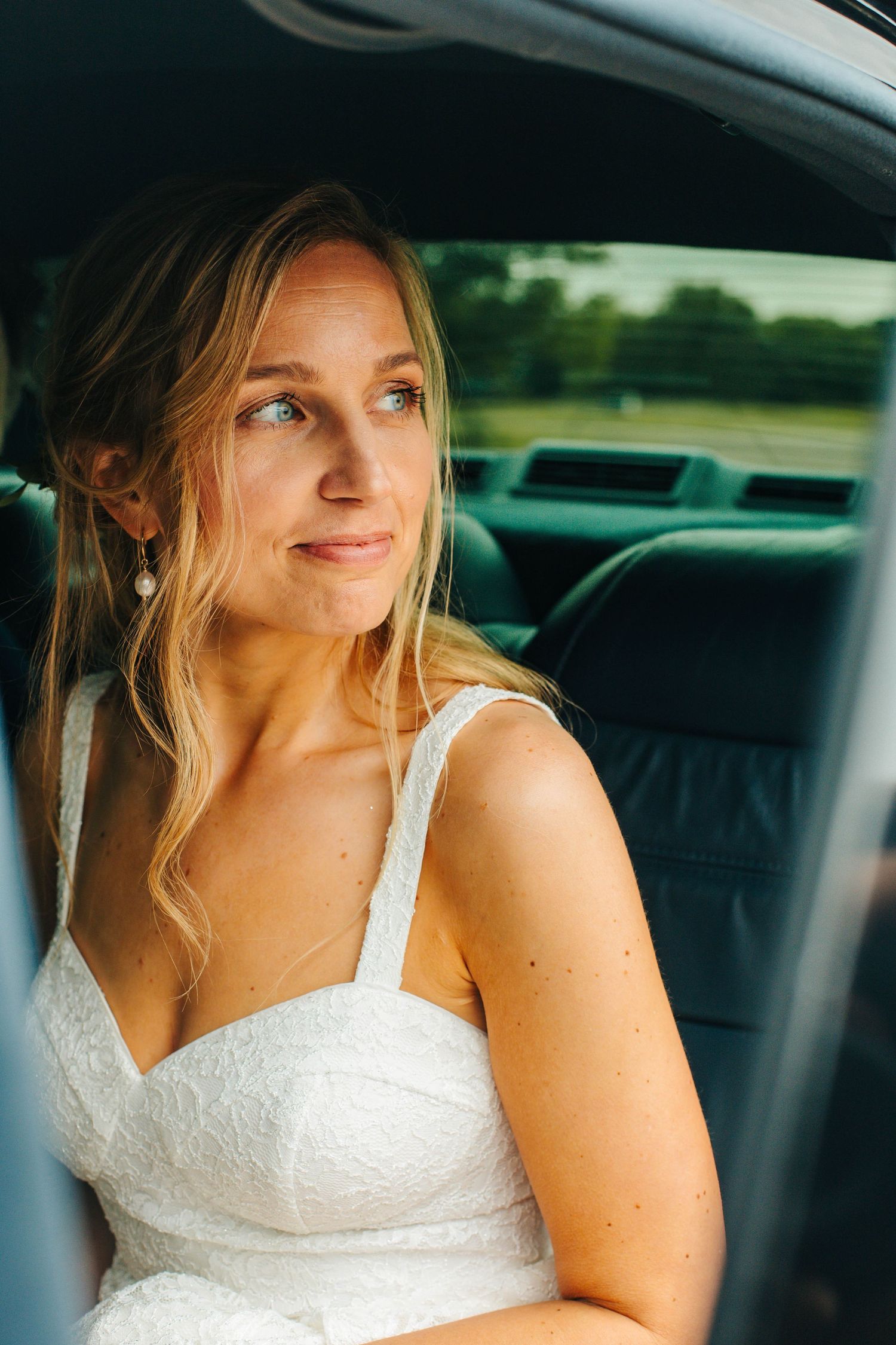 A smiling bride in a white dress sits in the front seat of a car on her wedding day.