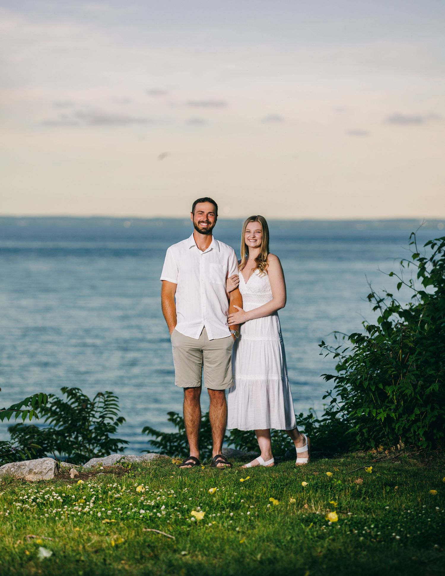 A couple stands on a grassy coastal bluff overlooking the ocean during golden hour.