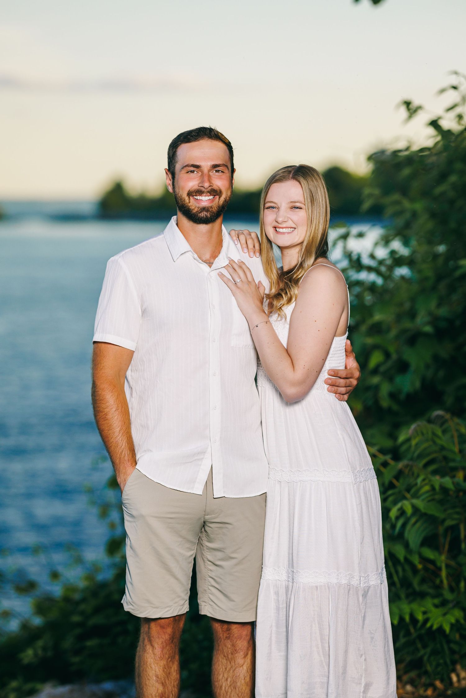 A couple wearing white outfits embraces while posing by the ocean during a romantic photoshoot at sunset.