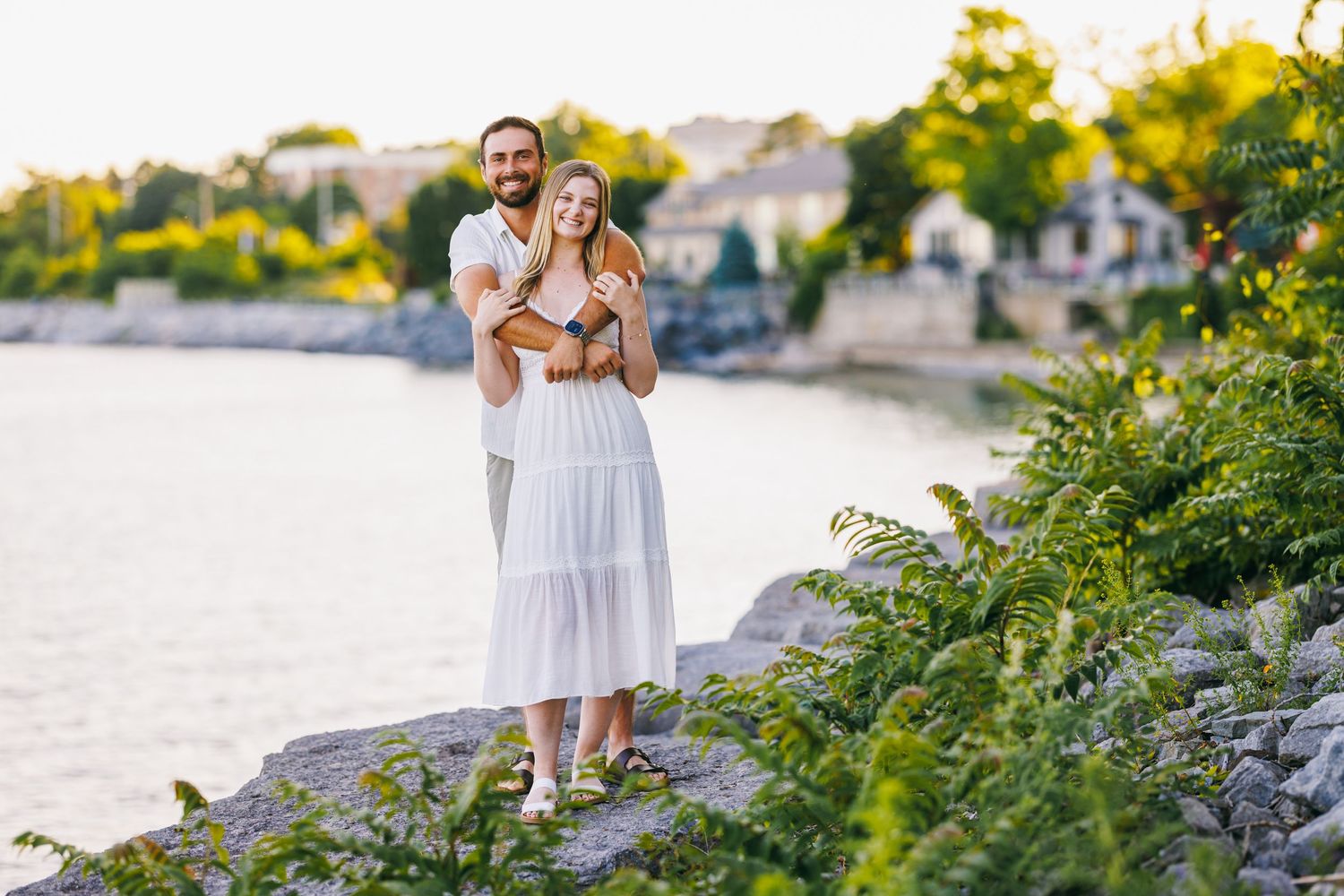 Two people in white outfits walking along a rocky beach shoreline at sunset near a residential area.