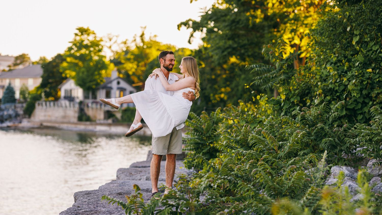 A romantic couple embrace on stone steps next to a tranquil waterfront at sunset in a lush garden setting.