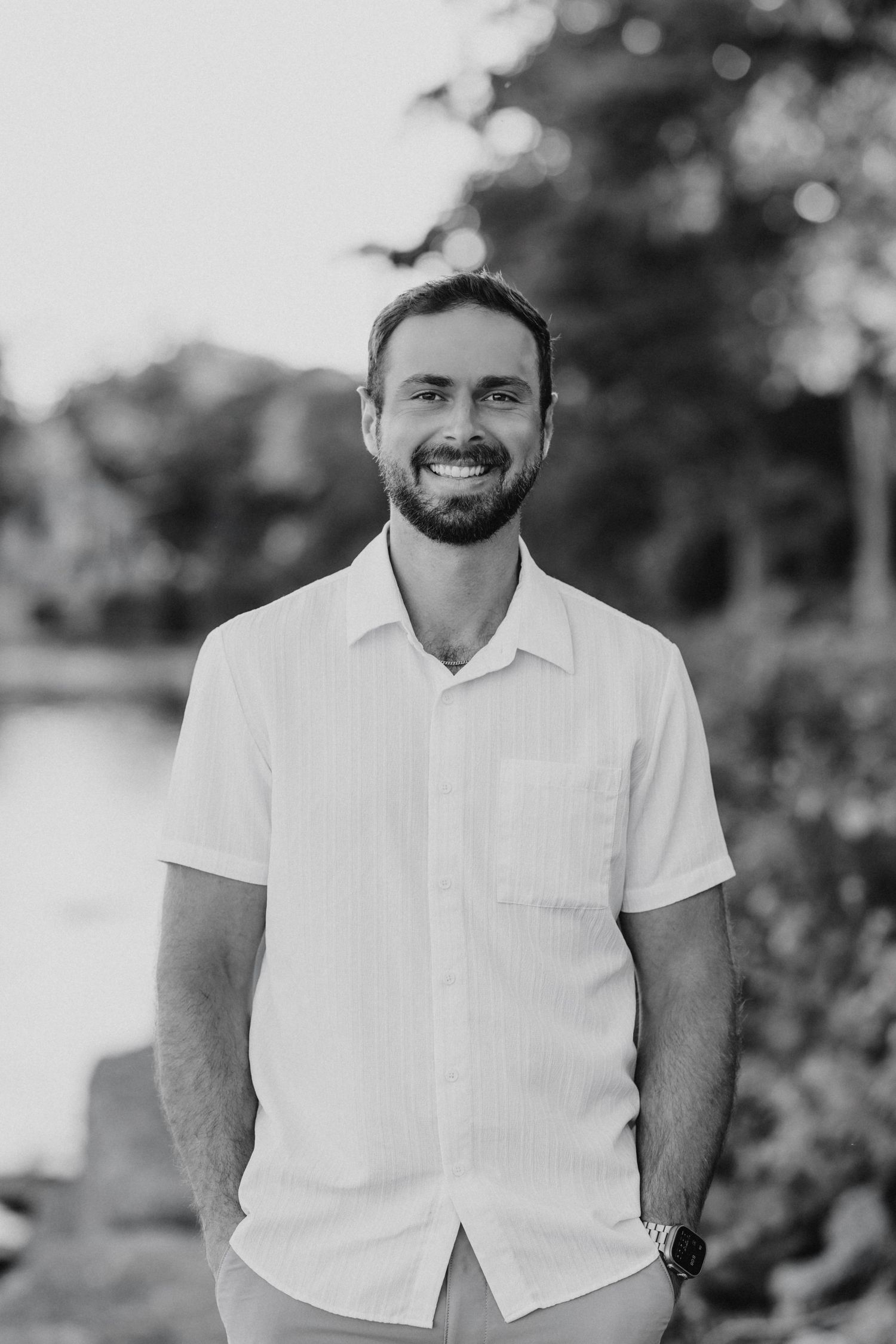 Black and white portrait of a bearded person wearing a white short-sleeve button-up shirt outdoors.