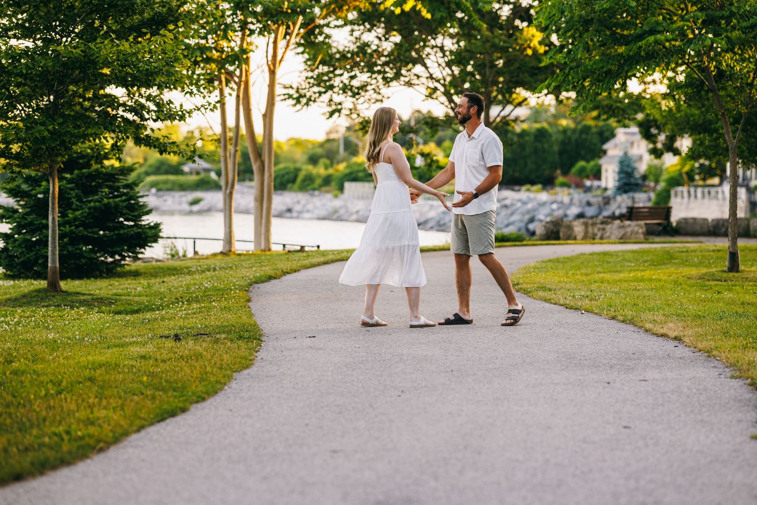 Two people in white and beige clothing walk together on a tree-lined path near a waterfront during golden hour.