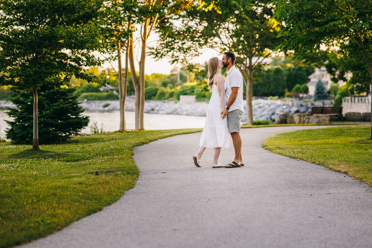 A couple walks hand in hand down a sunlit park path surrounded by green trees and grass.