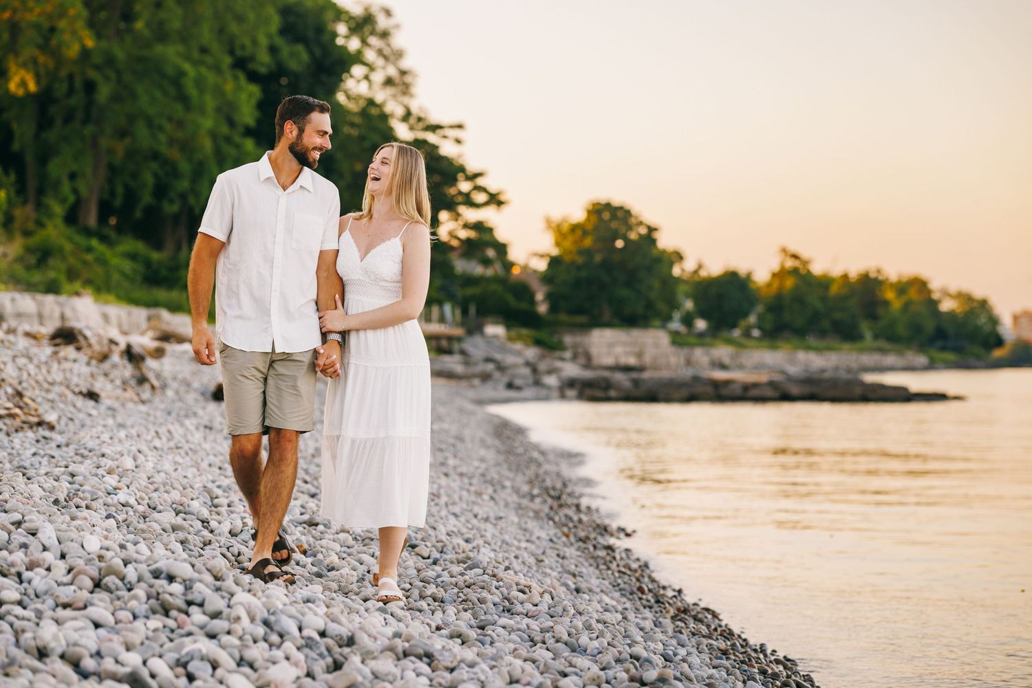 Couple walking together on a rocky beach at sunset wearing casual white summer outfits.