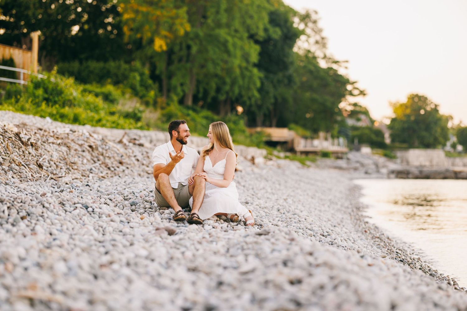 Couple walking hand in hand along a scenic shoreline with trees in background.
