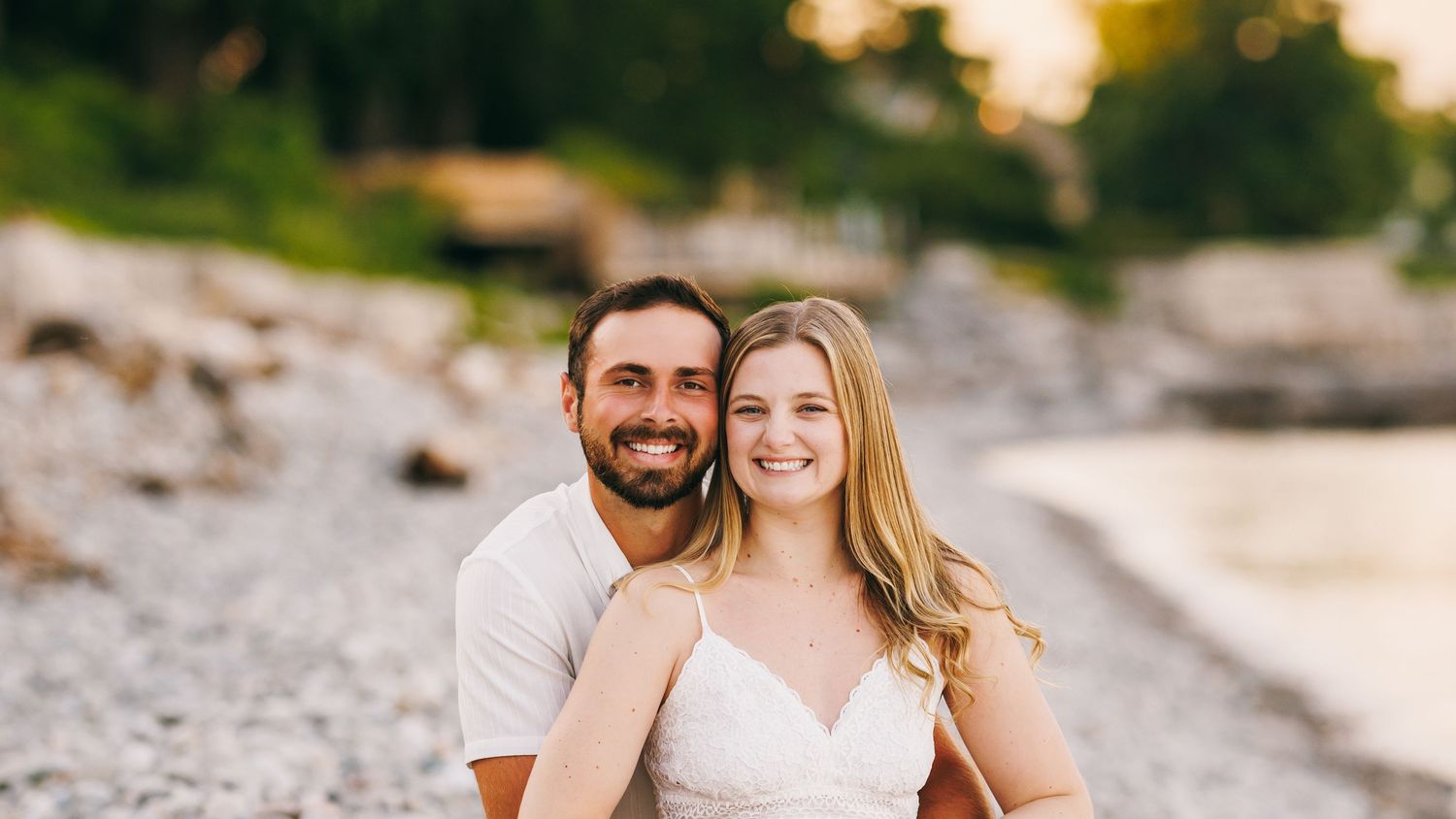 A happy couple embraces on a rocky beach during sunset with coastline and trees visible in the background.