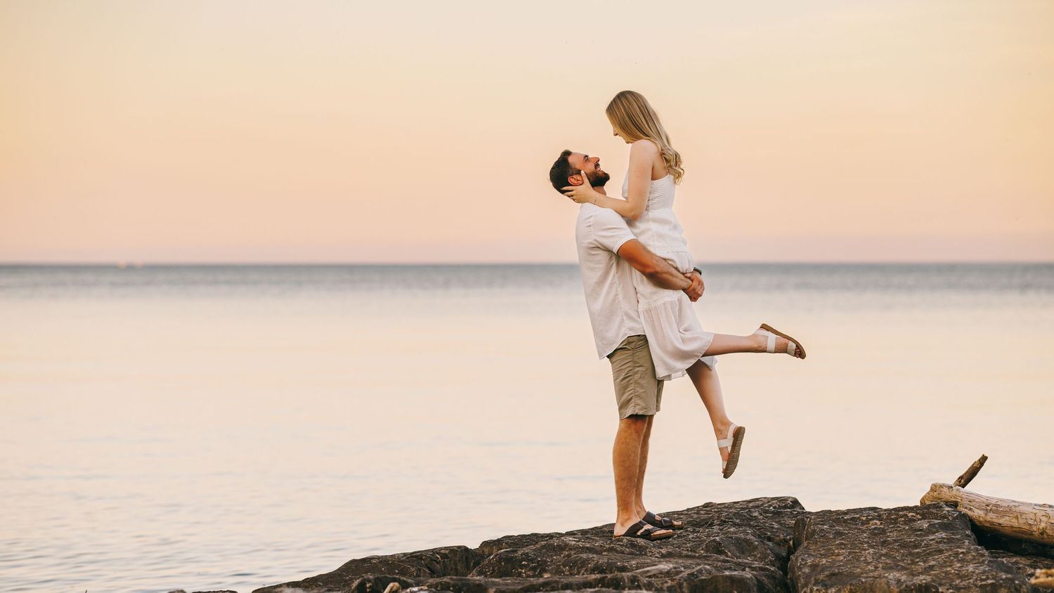 Silhouetted figures embrace on a rocky outcrop against a sunset sky over calm waters.