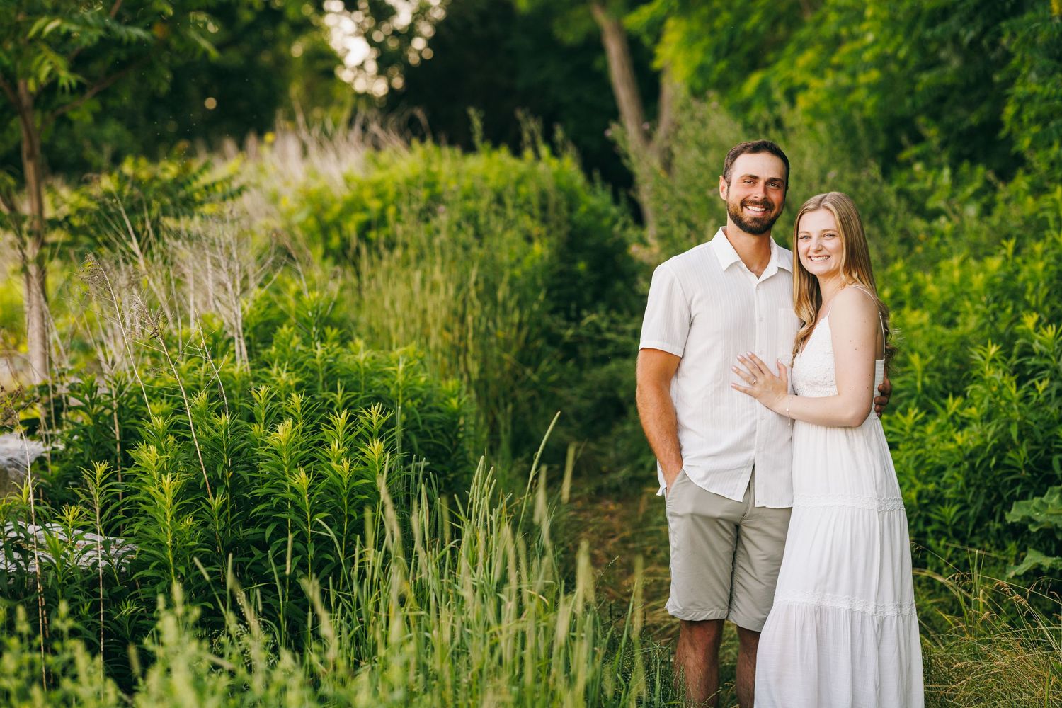 An affectionate couple in white outfits embraces while standing in a lush summer meadow with wildflowers and greenery.