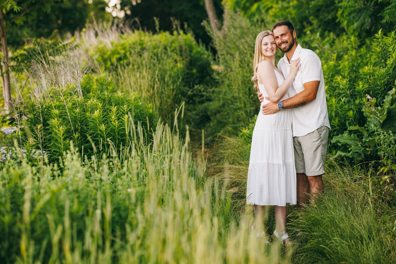 A romantic couple embraces in a lush green meadow during a summer evening photoshoot.