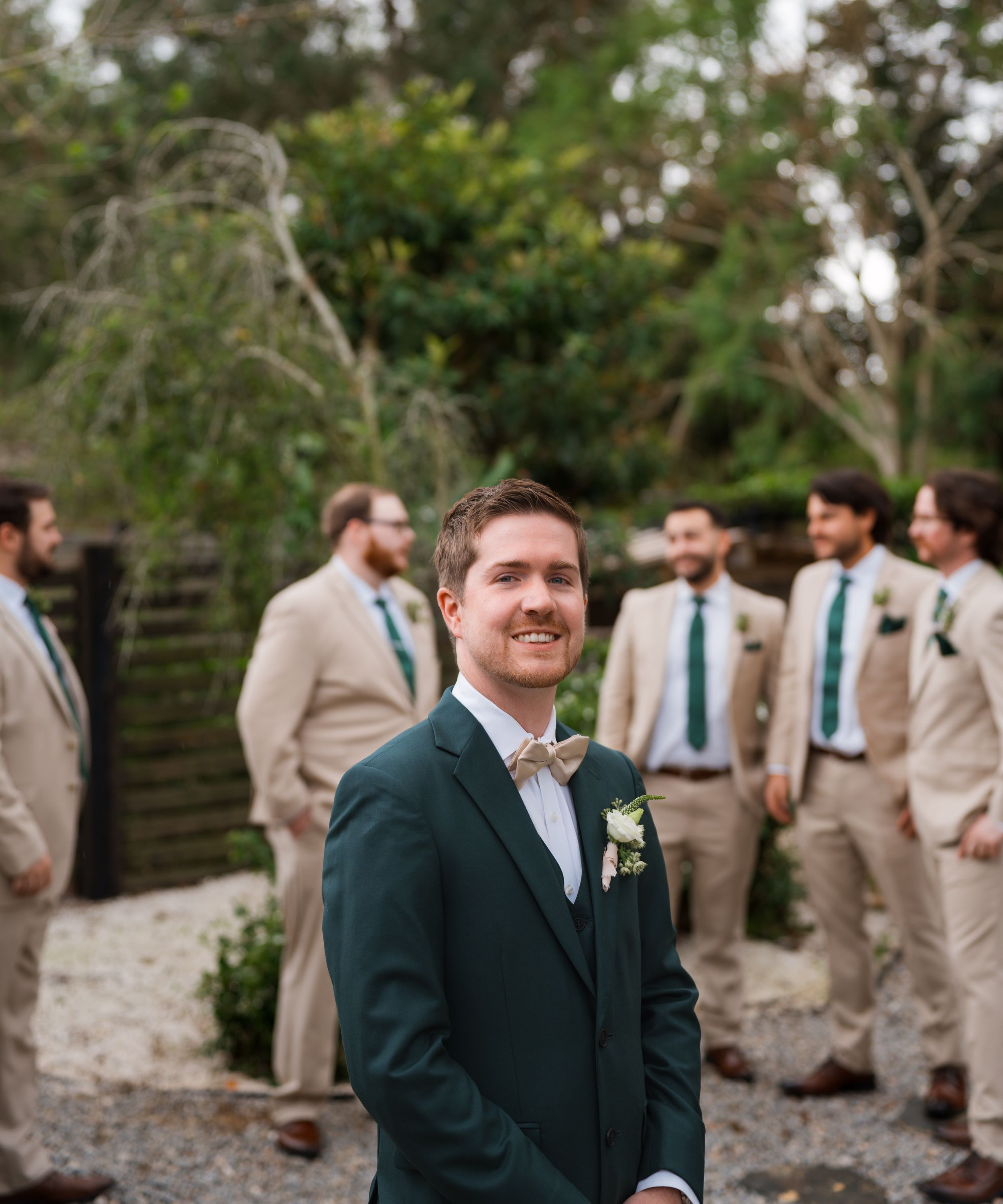 Wedding party gathers outdoors with groomsmen in tan suits and blue ties standing behind in a garden setting.