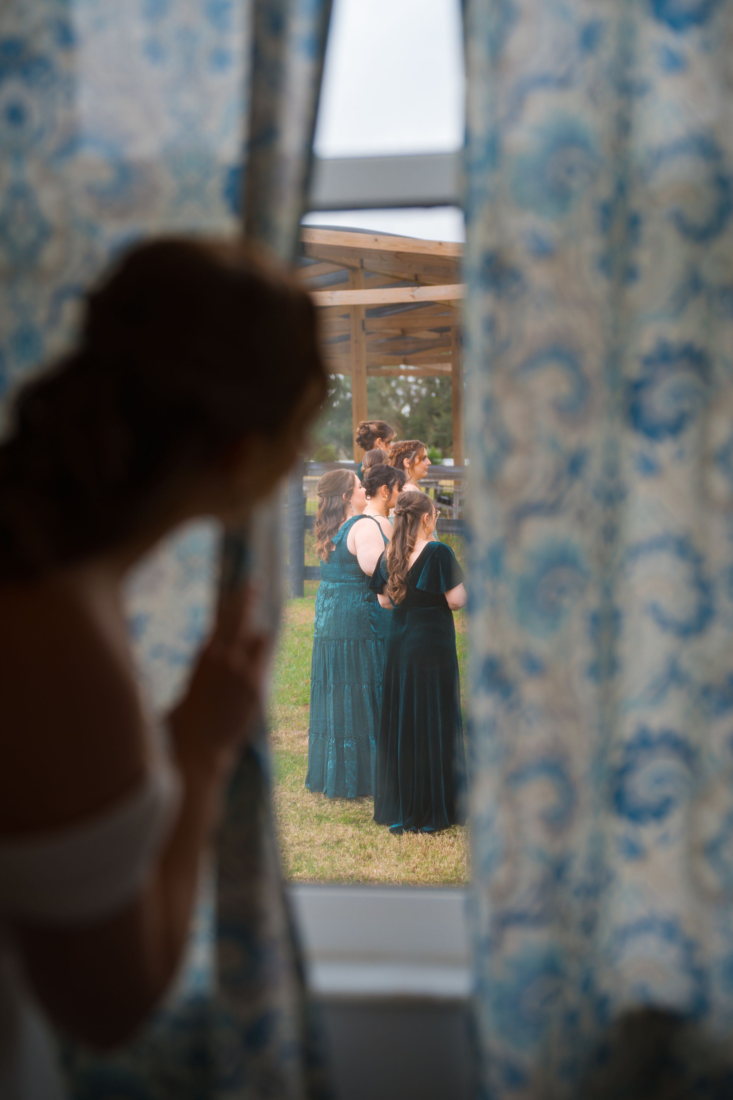 A peek through blue curtains reveals bridesmaids in teal dresses gathered outside on the lawn.