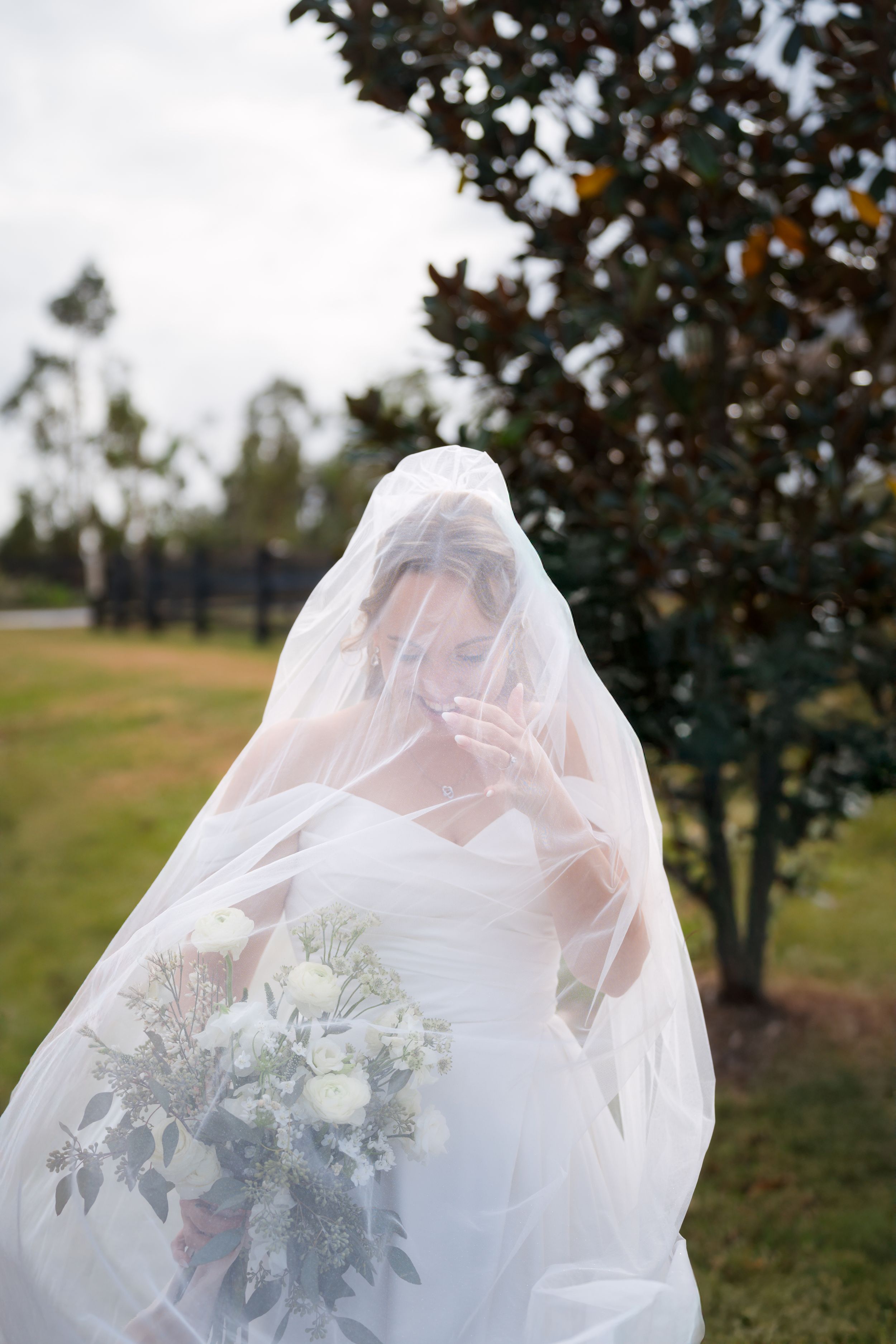 A bride in a white wedding dress stands beneath a flowing veil against a backdrop of trees at an outdoor ceremony.