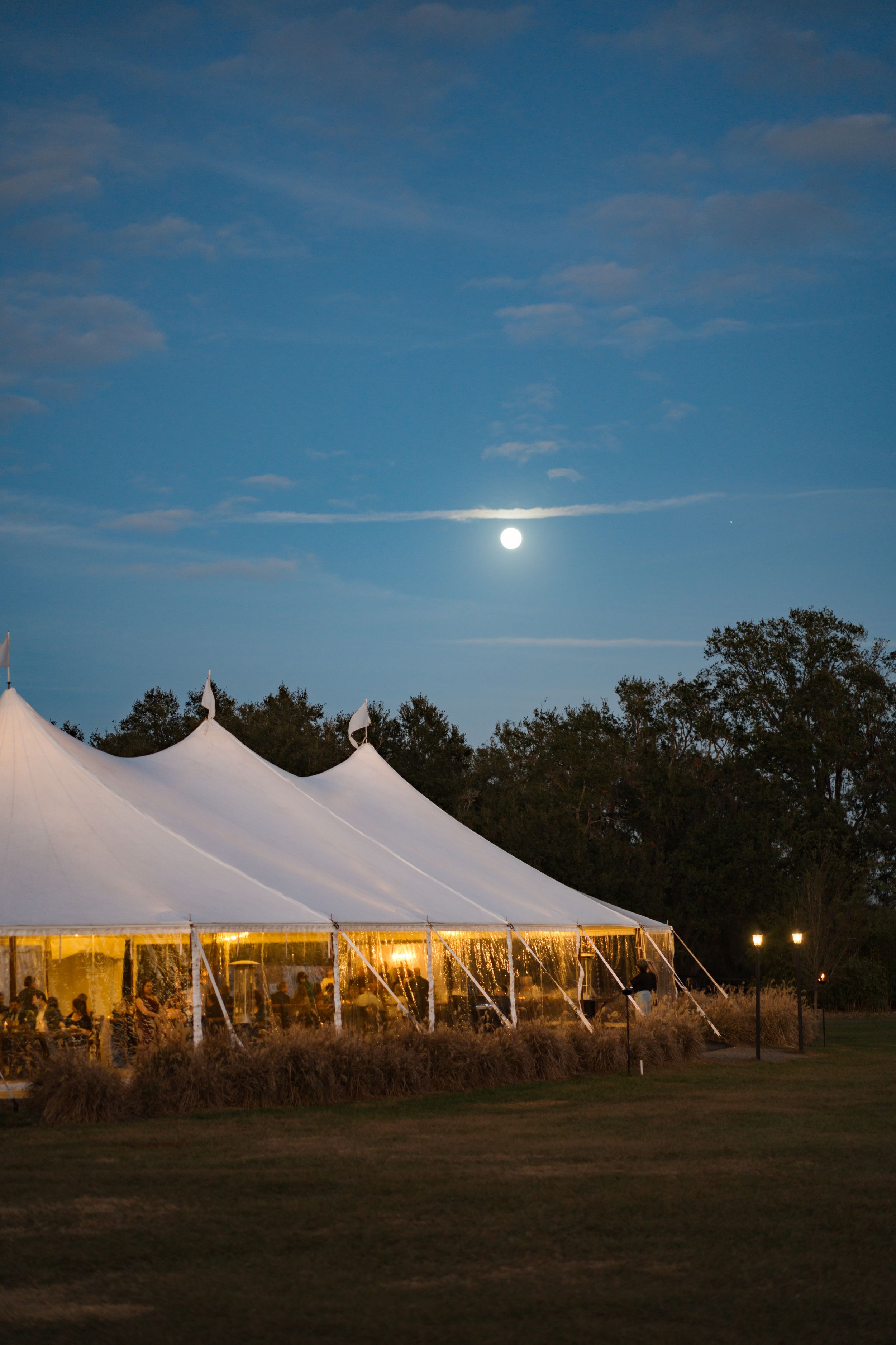 A large white wedding tent glows warmly under a full moon with trees silhouetted against a deep blue evening sky.