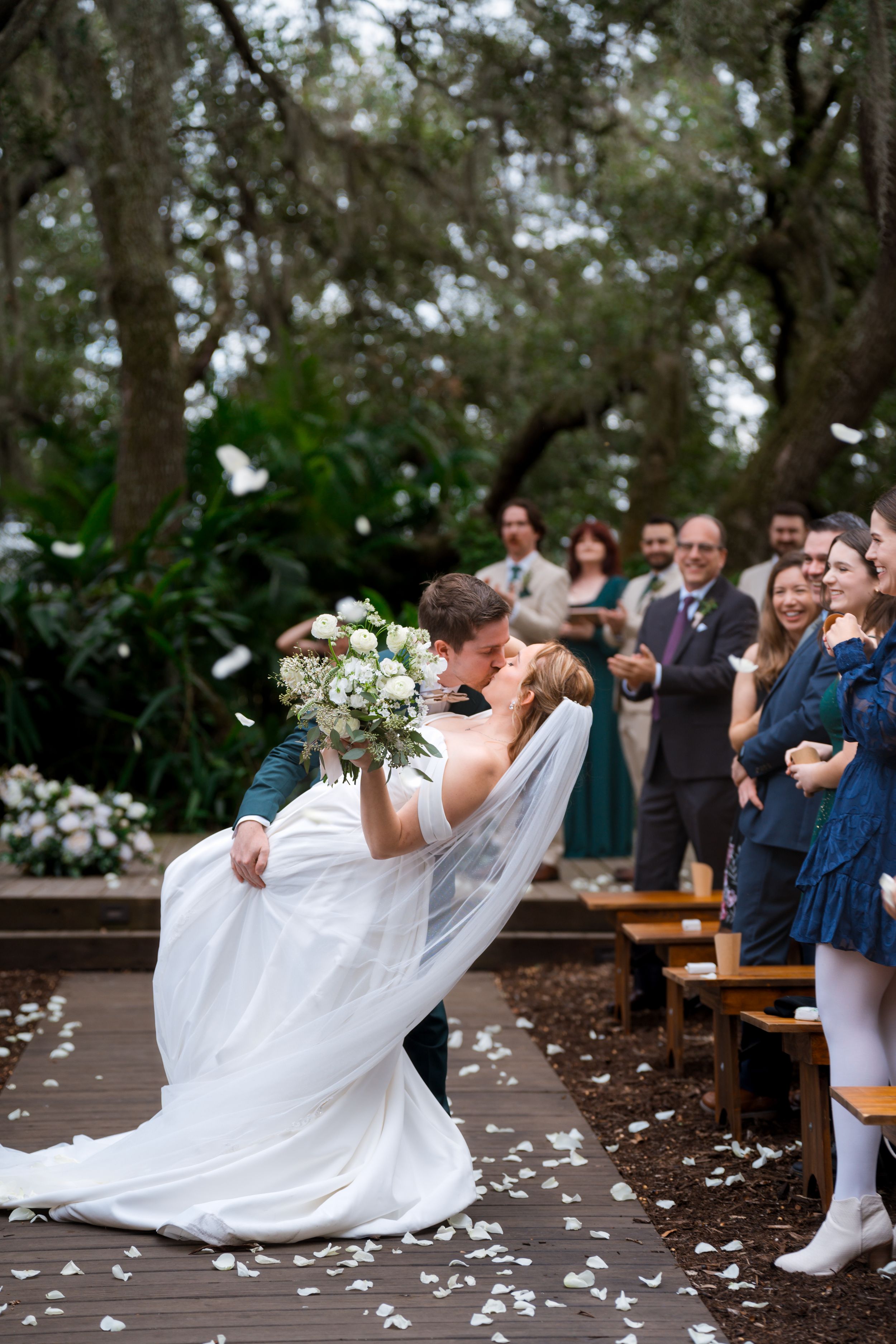 Wedding couple shares a romantic dip kiss at outdoor ceremony with seated guests and scattered flower petals.